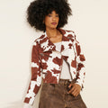 A model with a large black afro wears a brown and white cow-print moto jacket over a white top and brown denim jeans, posing against a plain off-white background.