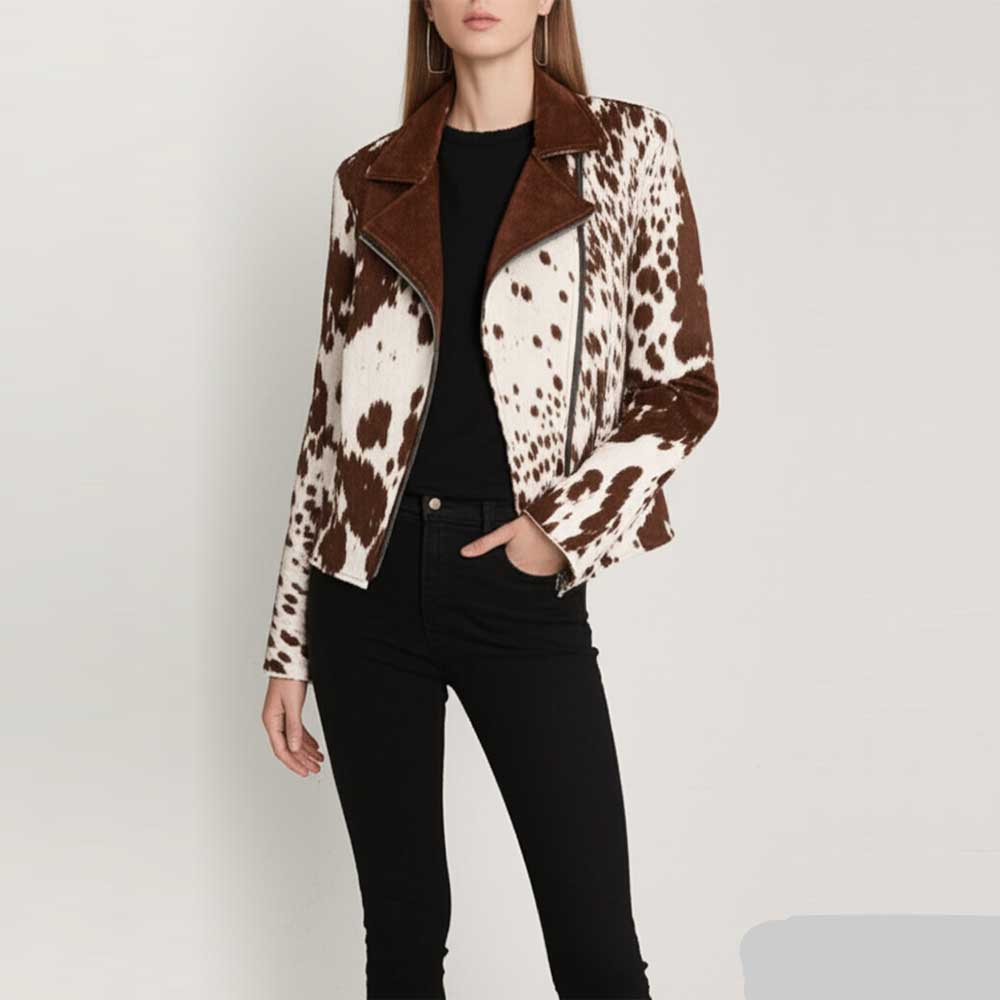 A woman models a cow-print moto jacket with brown suede-like lapels and collar, styled with a black top and black pants against a light gray studio background.