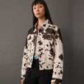 A model wearing a cropped cowhide jacket with a brown and cream cow-print pattern, styled with a cream top, black pants, and a turquoise belt buckle against a dark gray studio background.
