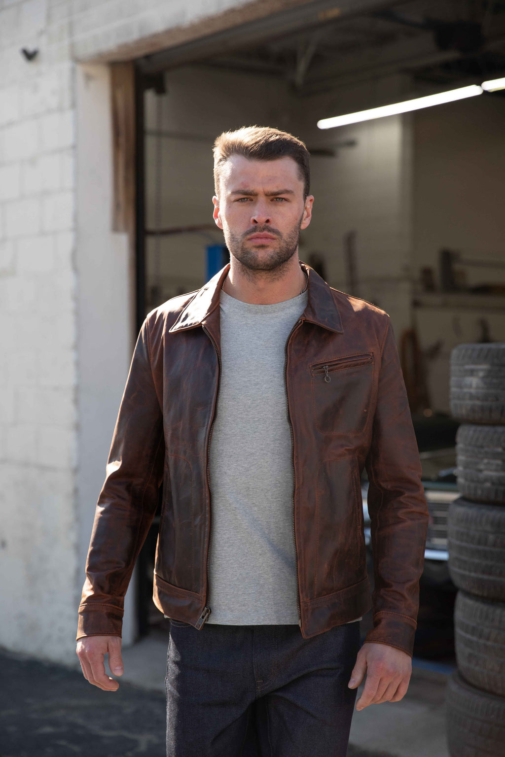A man wearing a distressed brown leather jacket over a light gray t-shirt and dark jeans, standing in front of an open garage door with tires stacked on the right.