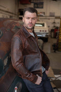 A man wearing a distressed brown leather jacket leans against a vintage car in a garage setting.
