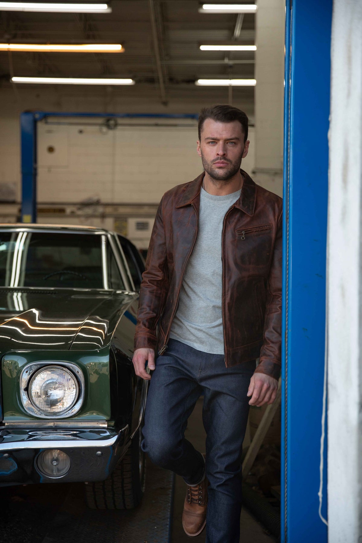 A man wearing a brown leather jacket, gray t-shirt, and dark jeans stands in a garage next to a green classic car.