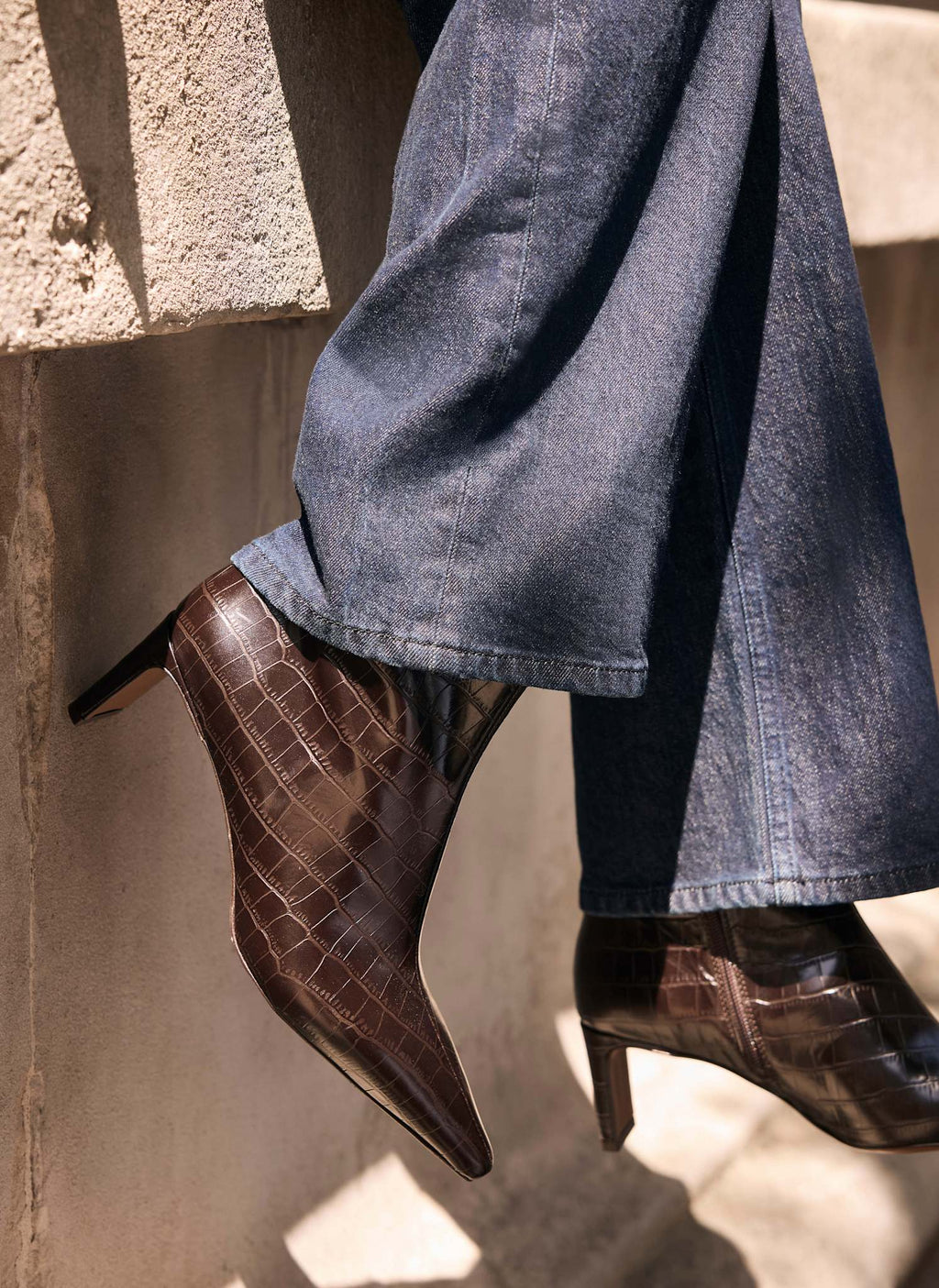 A close-up of a person's legs wearing dark brown crocodile-embossed ankle boots with pointed toes and mid-height heels, paired with wide-leg blue jeans, set against a textured concrete wall.
