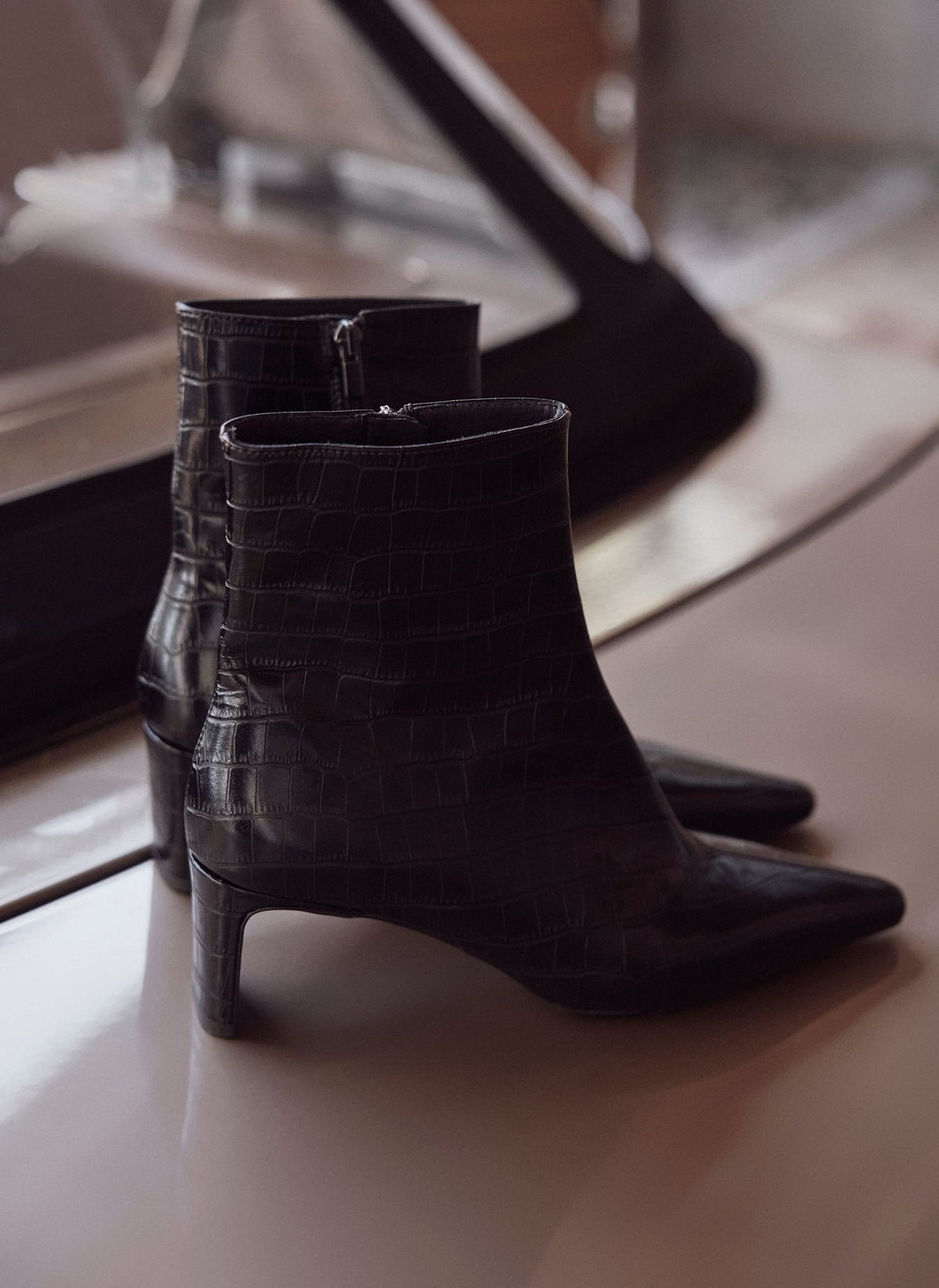 A pair of black pointed-toe ankle boots with a crocodile-embossed texture and kitten heels, resting on a reflective surface.