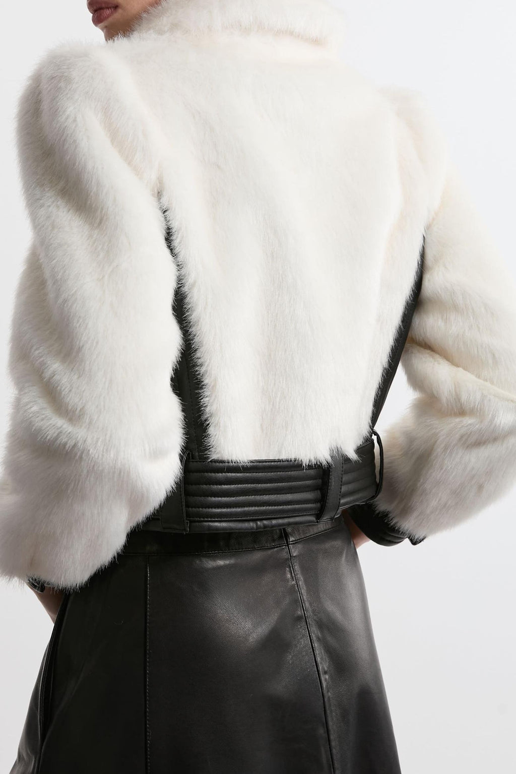 Close-up back view of a model wearing a white fur cropped jacket with black shoulder panels and a wide black belt, paired with a black leather skirt.