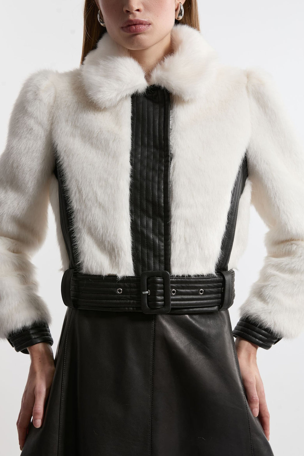 A close-up of a model wearing a white fur jacket with black faux-leather trim, a black pleated front panel, and a wide black belt.