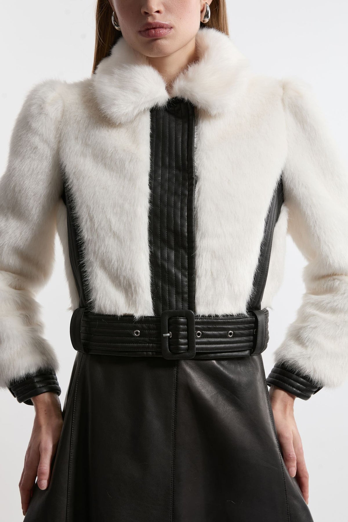 A close-up of a model wearing a white fur jacket with black faux-leather trim, a black pleated front panel, and a wide black belt.