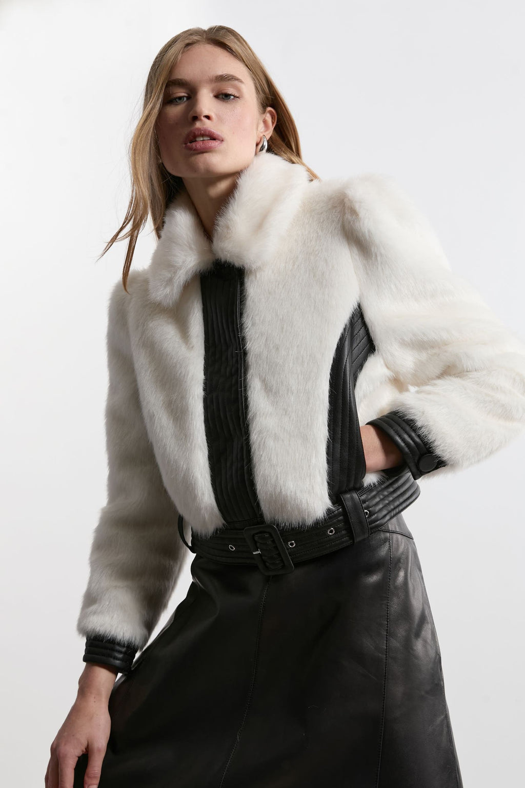 A model wearing a white fur jacket with black ribbed trim and a black leather skirt, photographed against a plain light gray studio background.
