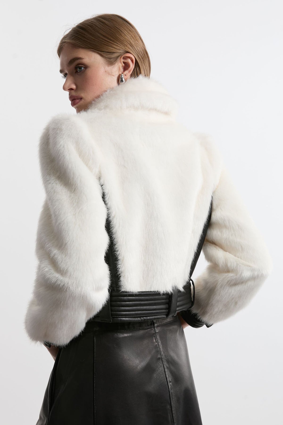A model shown from the back wearing a white fur cropped jacket with a high collar and black leather skirt against a plain white background.