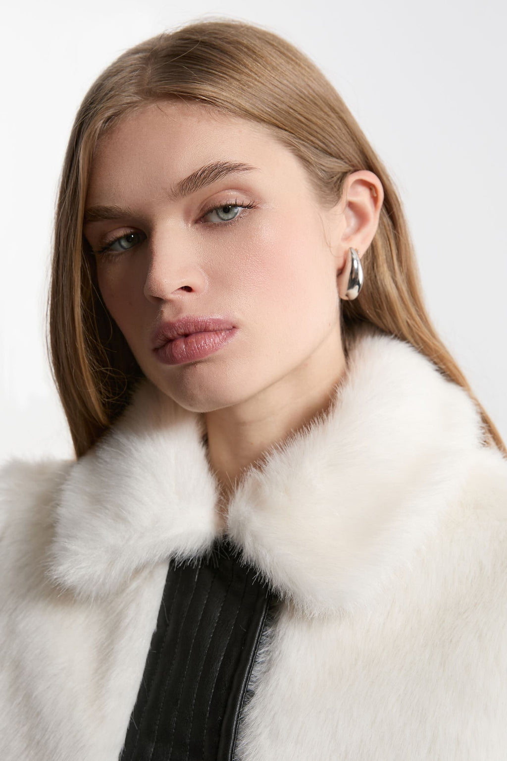 A close-up portrait of a woman wearing a luxurious white fur collar with a black ribbed garment underneath, set against a plain light gray background.