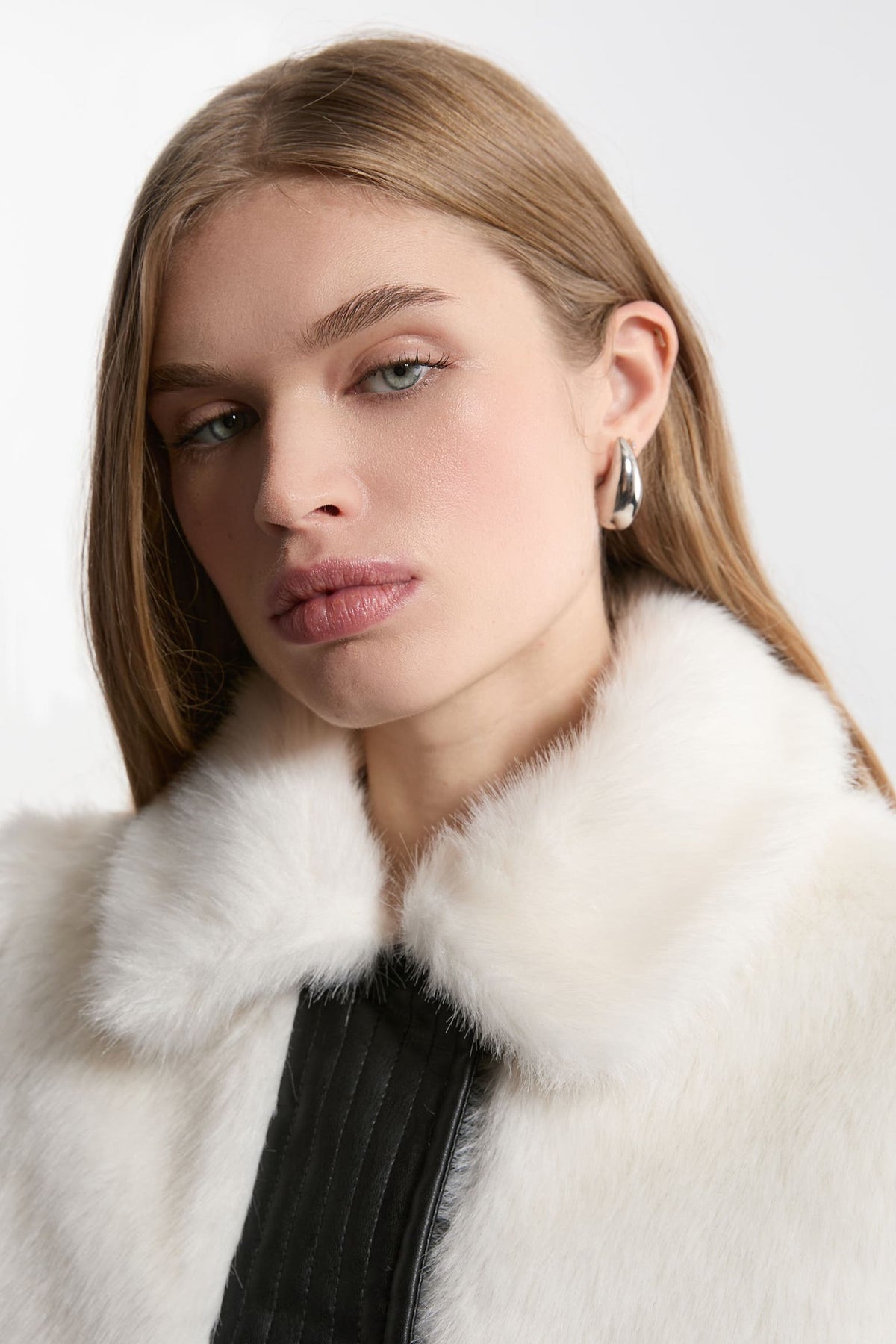 A close-up portrait of a woman wearing a luxurious white fur collar with a black ribbed garment underneath, set against a plain light gray background.