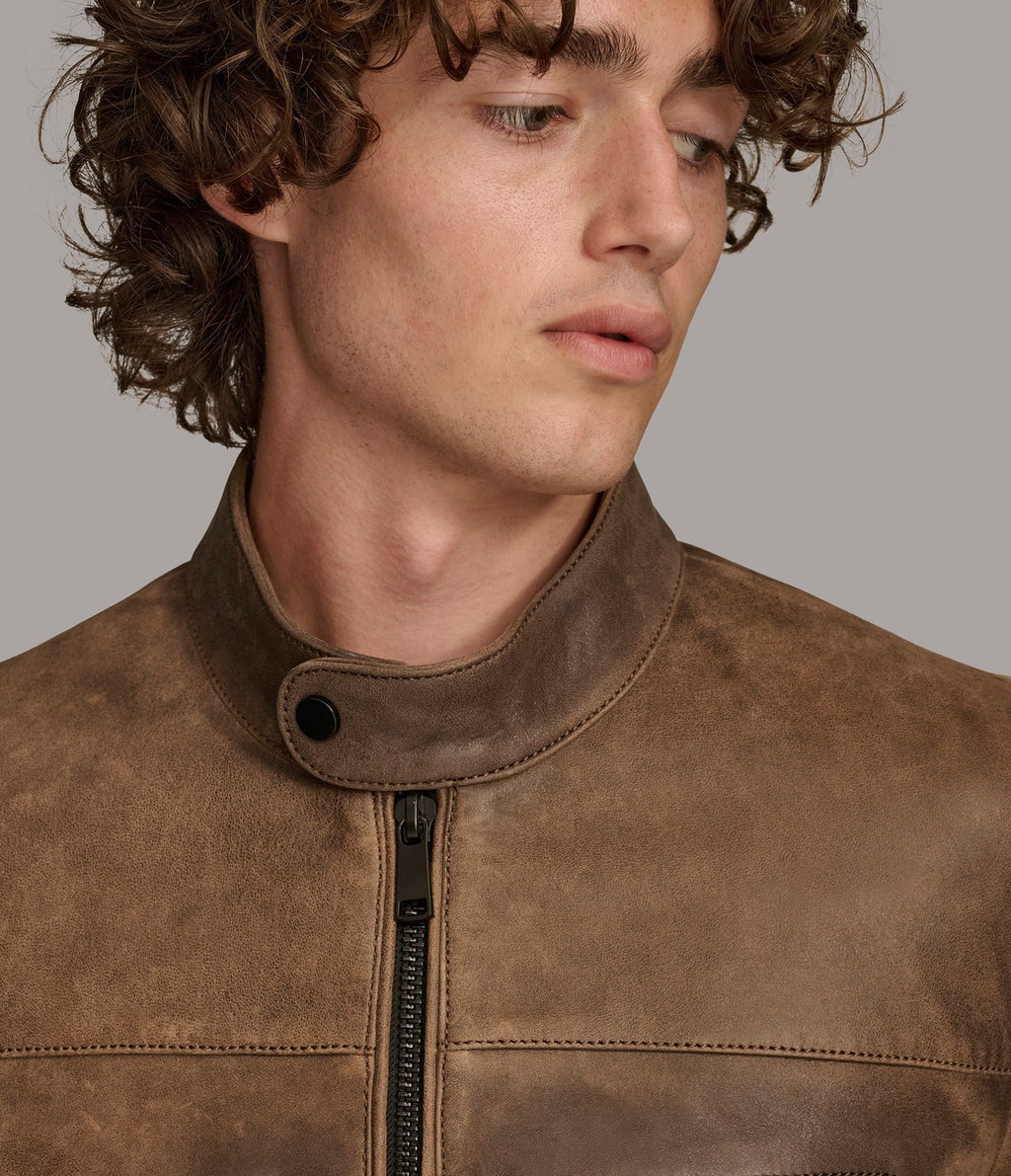 Close-up of a man wearing a brown suede or nubuck motorcycle jacket with a high collar and black zipper, against a solid gray background.