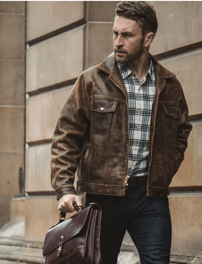 A man wearing a brown distressed leather jacket over a plaid shirt and dark jeans, holding a dark brown leather briefcase against a stone building background.