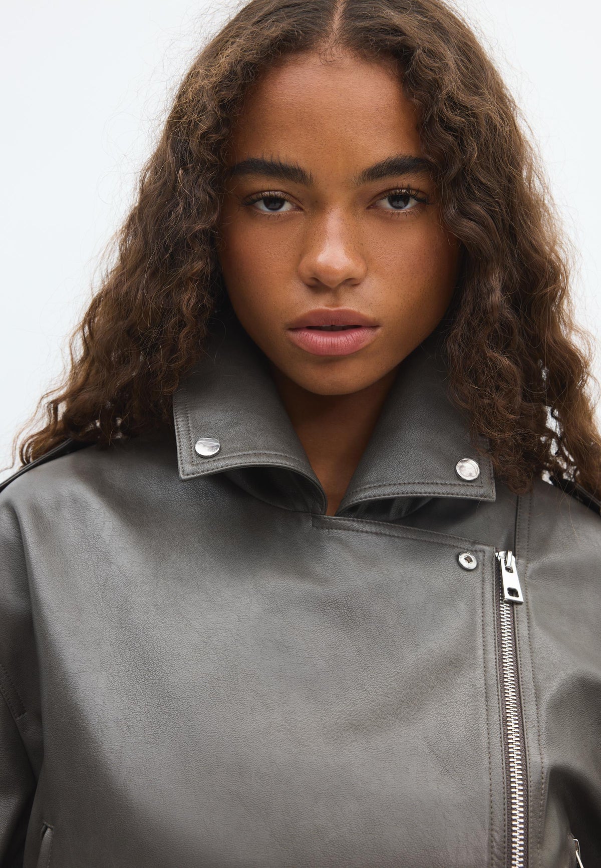 A close-up of a model wearing a dark gray faux-leather biker jacket with an asymmetrical zipper and silver snap details on the collar.