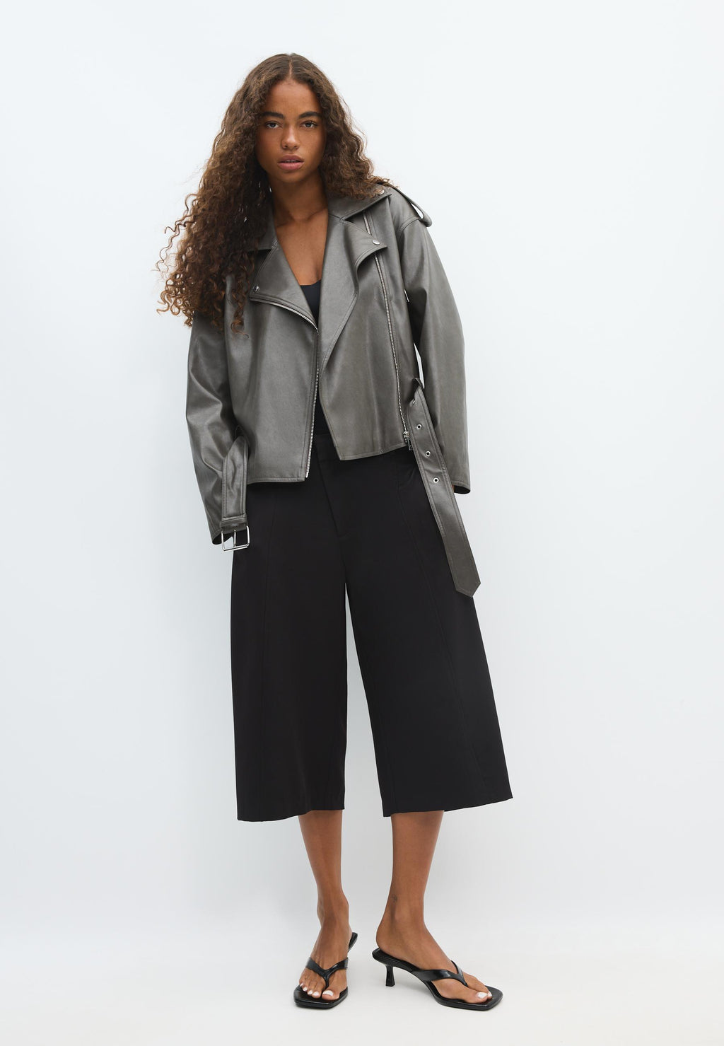 A model stands against a plain white studio background wearing a metallic silver moto jacket, black cropped wide-leg pants, and black heeled sandals.