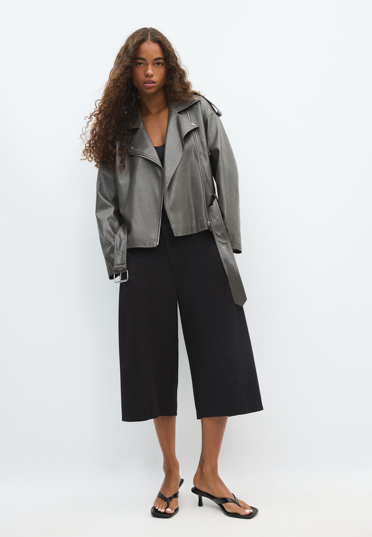 A model stands against a plain white studio background wearing a metallic silver moto jacket, black cropped wide-leg pants, and black heeled sandals.