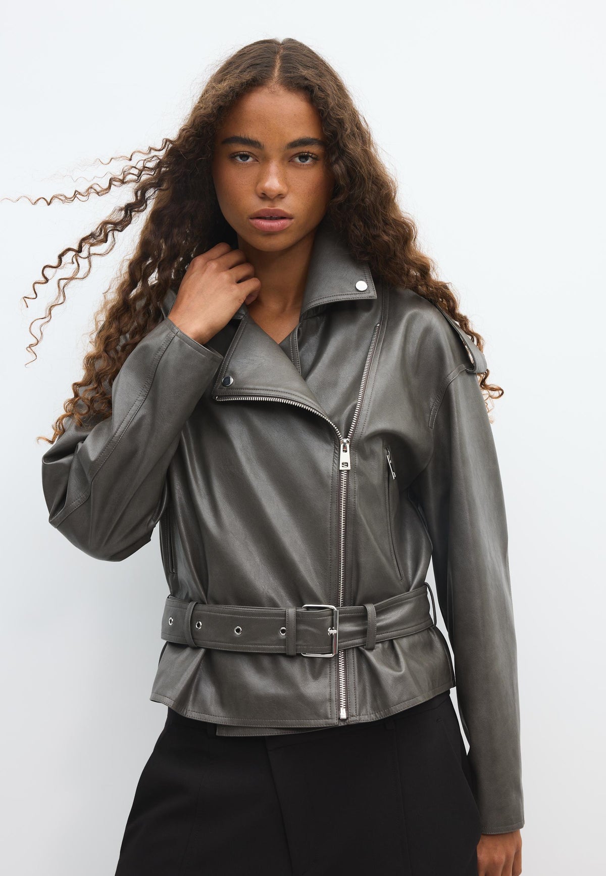 A woman modeling a dark gray faux-leather moto jacket with a belted waist and silver zipper details, paired with black pants, against a plain light gray studio background.