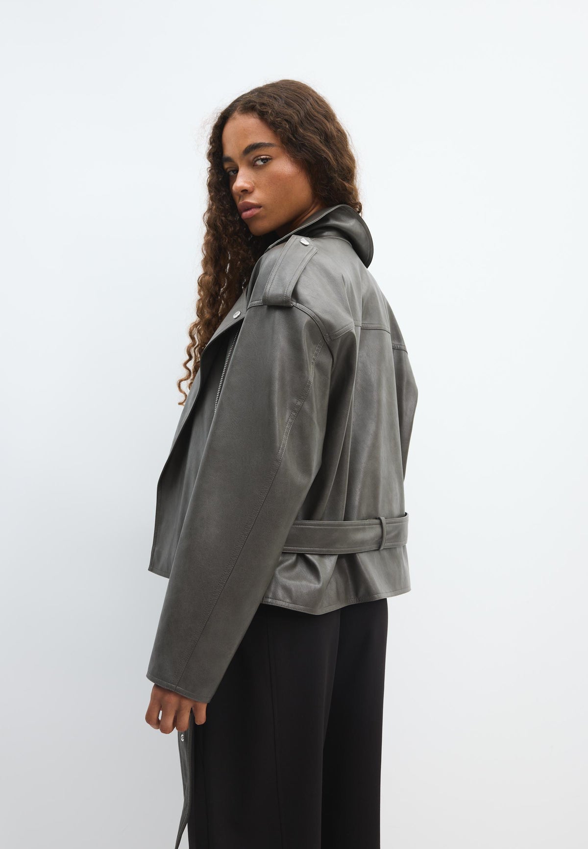 A woman modeling a cropped gray leather jacket with a belted waist, shown from a side profile against a plain light gray studio background.