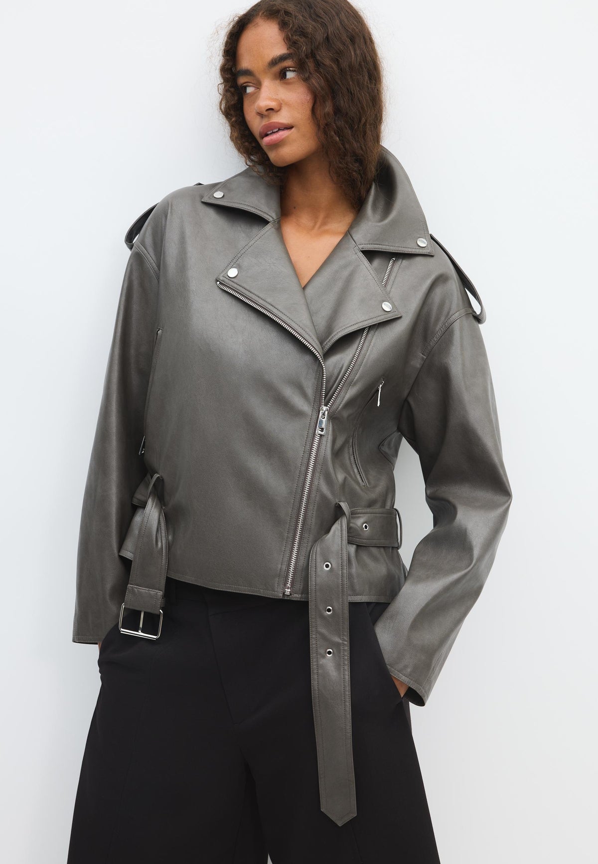 A woman wearing a cropped, dark gray faux-leather moto jacket with a belted waist and silver hardware, paired with black pants, against a plain white studio background.