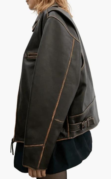 A side-profile view of a model wearing a dark brown leather jacket with contrasting tan stitching, paired with a dark skirt, against a plain white background.