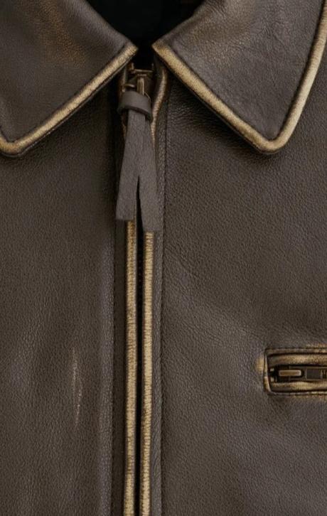 Close-up of a dark brown leather jacket with a collar, showing a dark brown zipper with a pull tab and a contrasting light tan stitched edge.