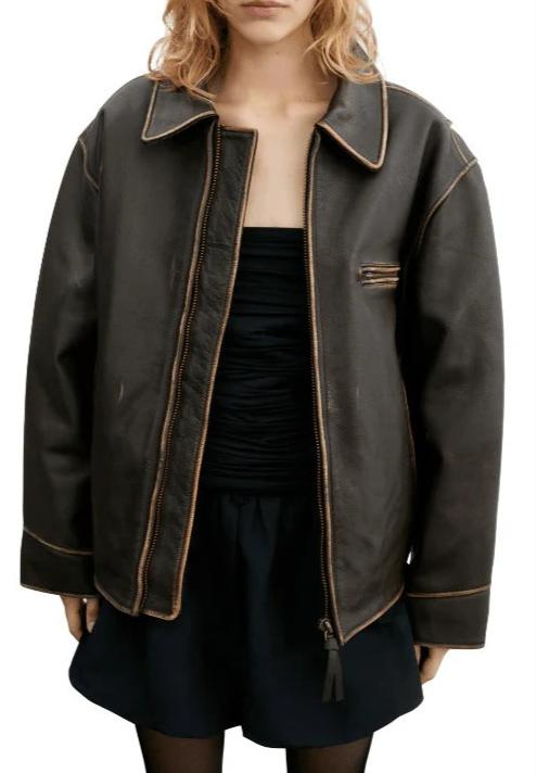 A model wearing an oversized dark brown leather jacket with tan contrast stitching, paired with a black top and skirt, against a plain white background.