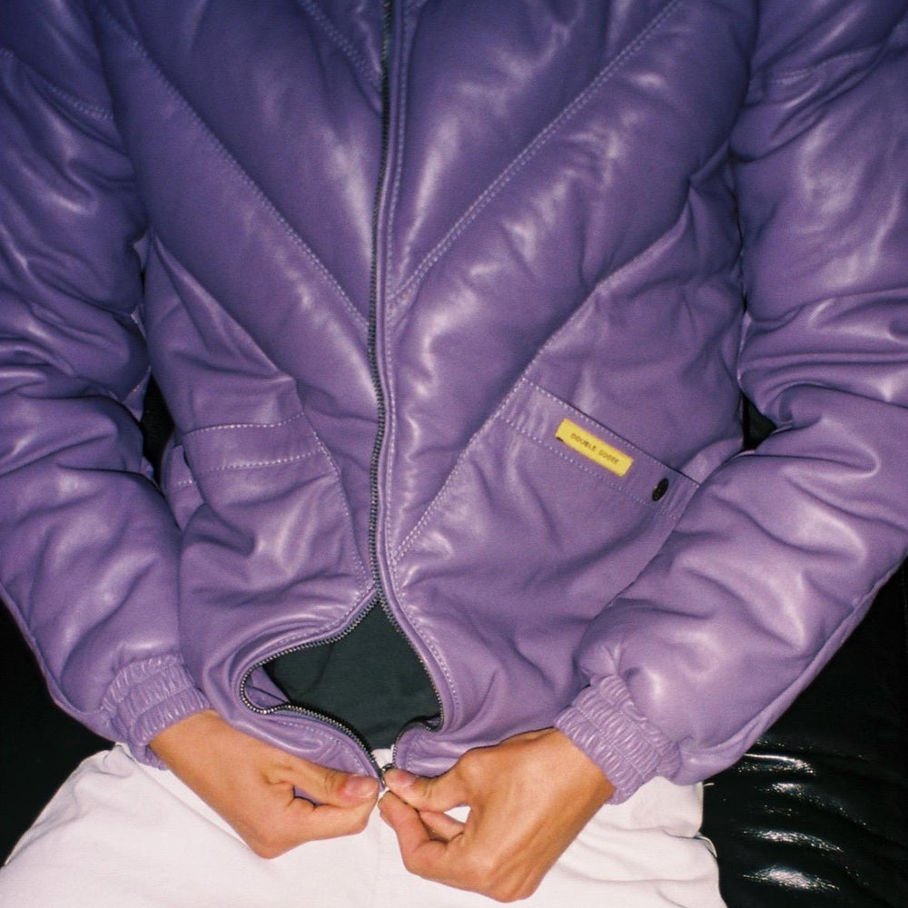 A close-up of a person wearing a shiny lavender-purple quilted jacket, pulling the zipper down with both hands. The jacket has a small yellow brand label on the pocket and is paired with white pants.