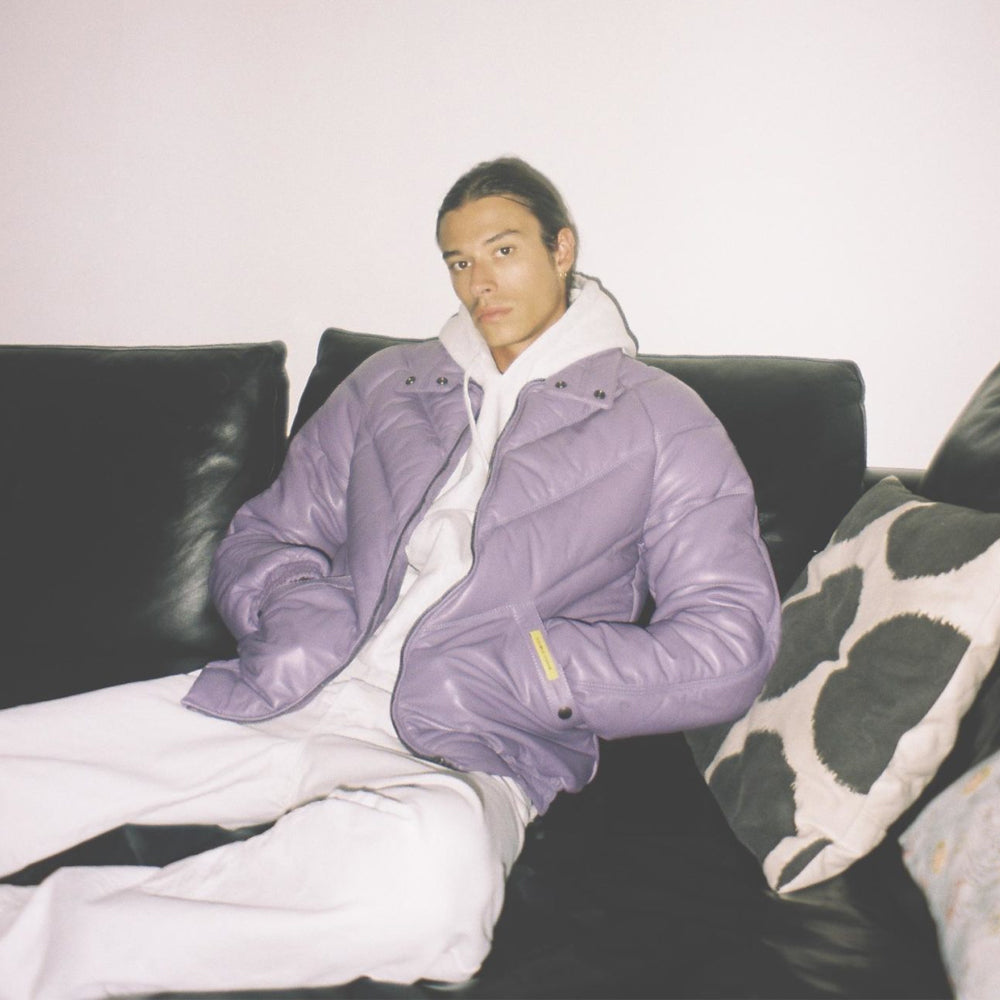 A man wearing a shiny lavender puffer jacket over a white hoodie and white sweatpants, sitting on a black leather couch with patterned pillows.
