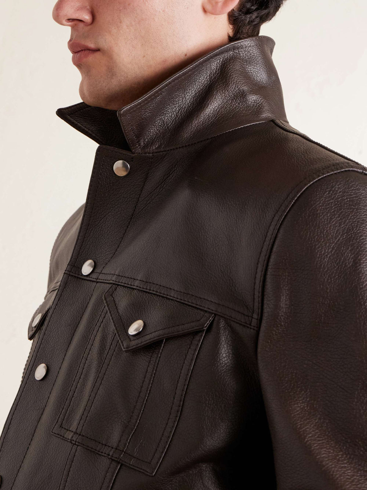 Close-up of a person wearing a dark brown leather jacket with a high collar and silver snap buttons, against a plain light background.