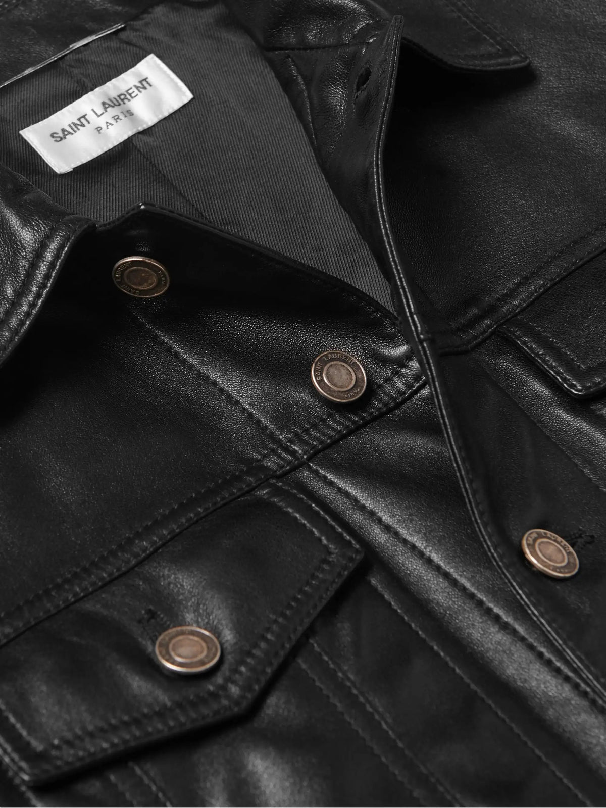 Close-up of a black leather jacket with a white Saint Laurent Paris brand label visible inside the collar, featuring bronze-toned metal snap buttons and a textured leather surface.