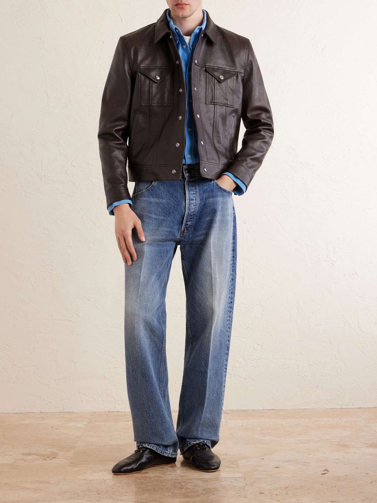 A man wearing a dark brown leather jacket over a blue button-down shirt, paired with blue jeans and black leather shoes, standing against a textured beige wall.