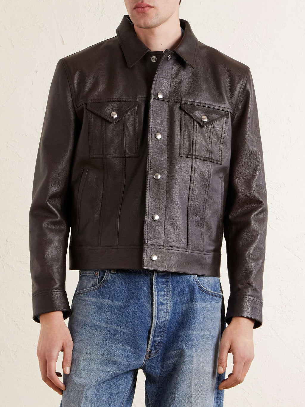 A man wearing a dark brown leather jacket with silver snap buttons and blue jeans, shown from the chin down against a light textured wall.