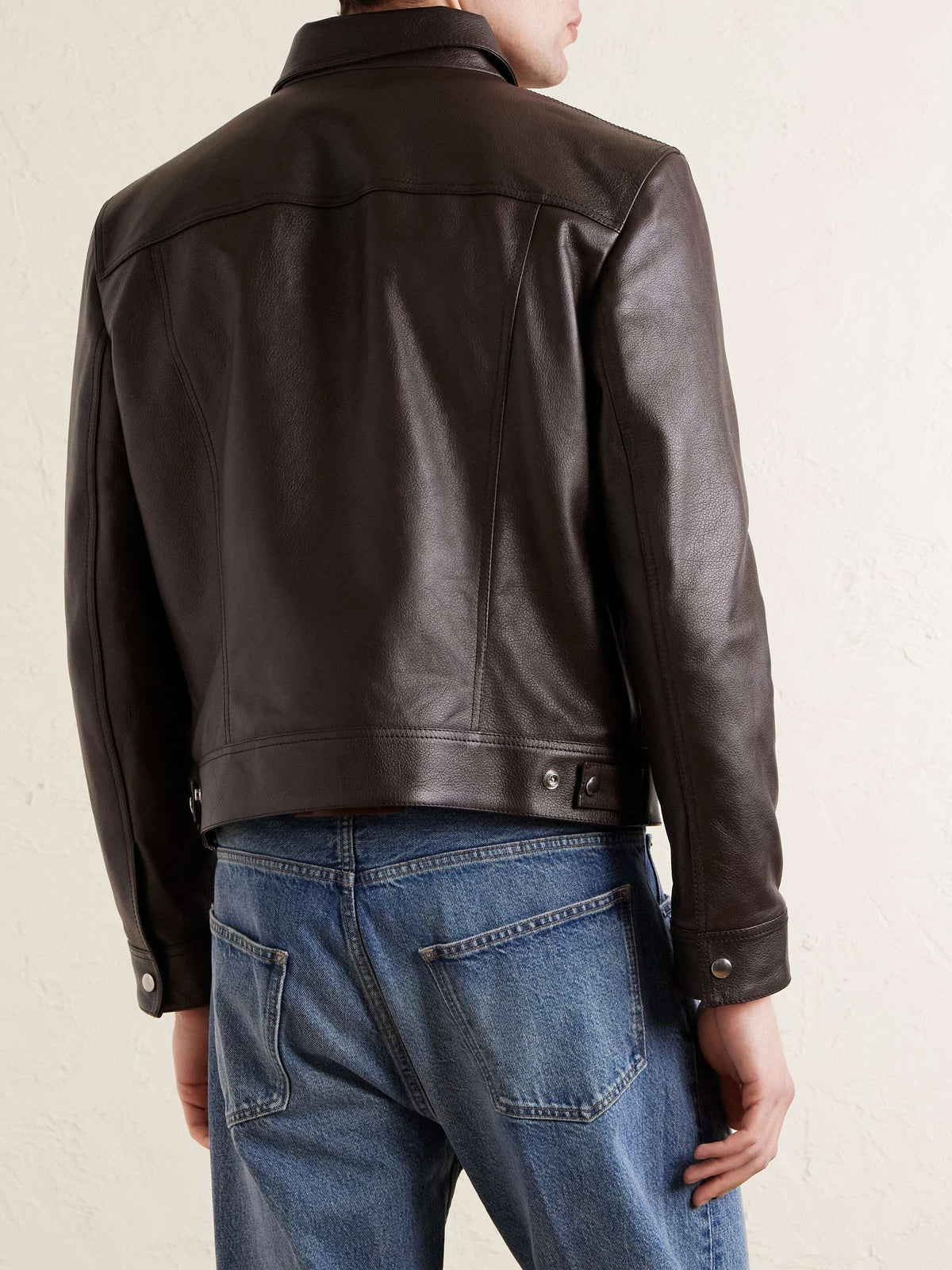 A man wearing a dark brown leather jacket, shown from the back, paired with blue jeans against a light textured wall.