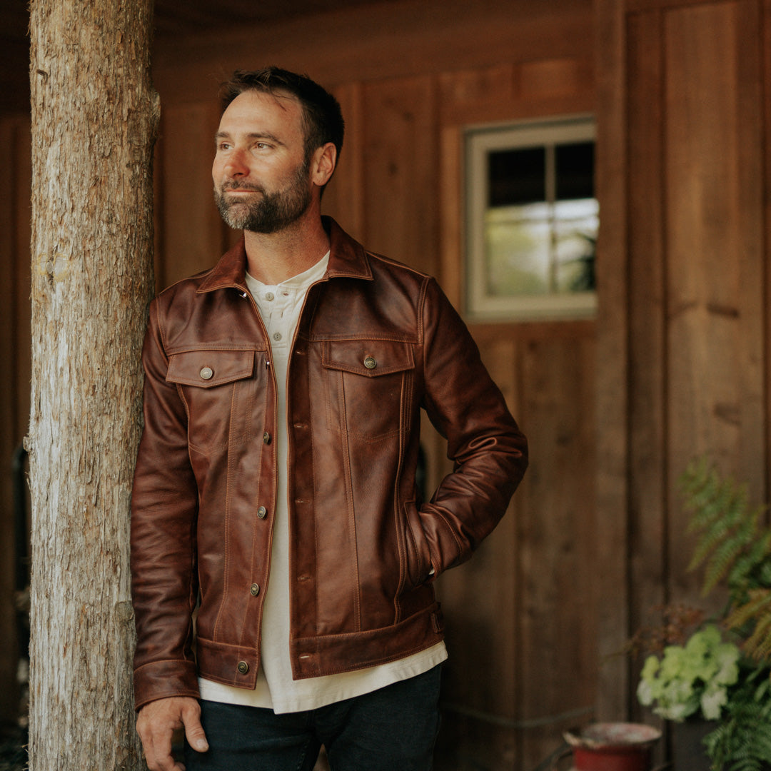 Man wearing a brown leather jacket standing against a wooden wall.