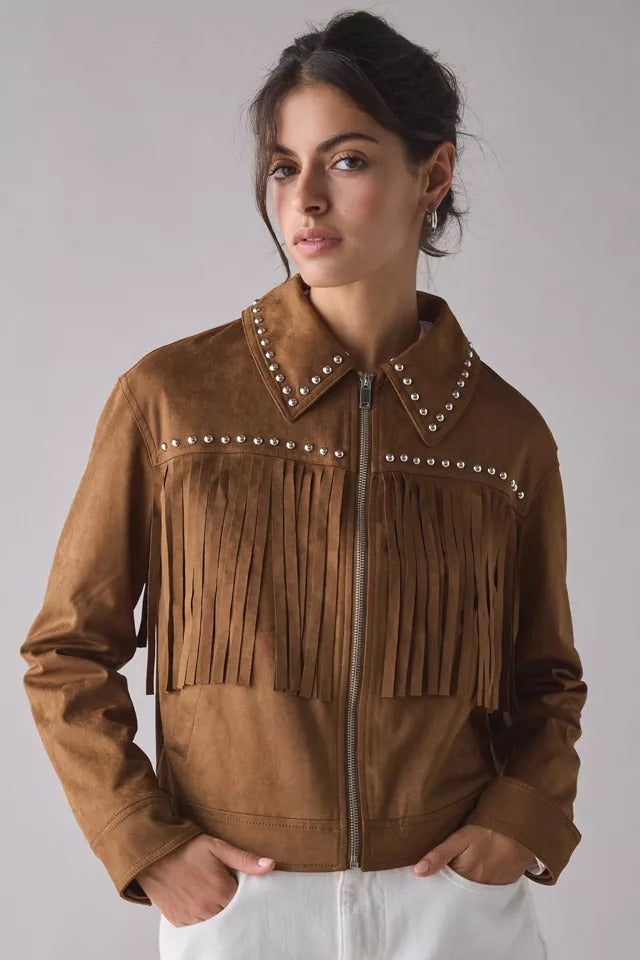 A woman wearing a brown suede fringe jacket with silver stud embellishments on the collar and chest, paired with white pants, photographed against a neutral gray studio background.