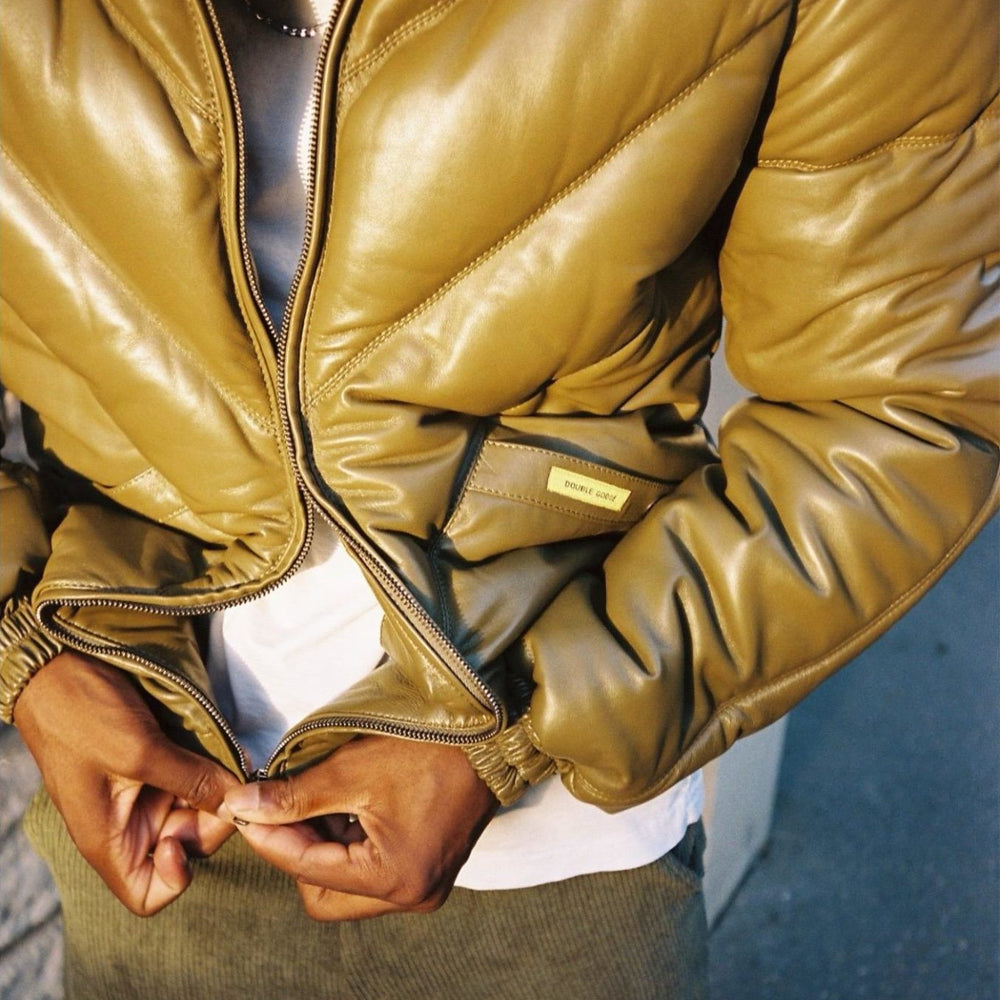 A close-up of a person wearing a shiny olive-brown leather puffer jacket, zipping it up over a white shirt. The jacket has a small rectangular brand patch on the pocket