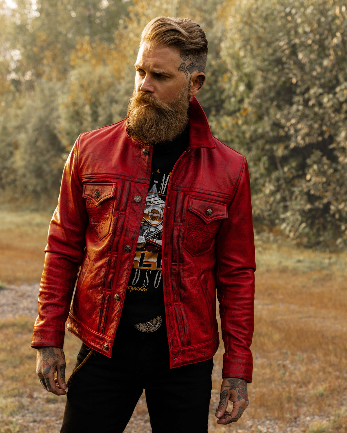 A man with a long beard and tattoos wearing a red leather jacket over a graphic t-shirt and black pants, standing outdoors in a wooded area.