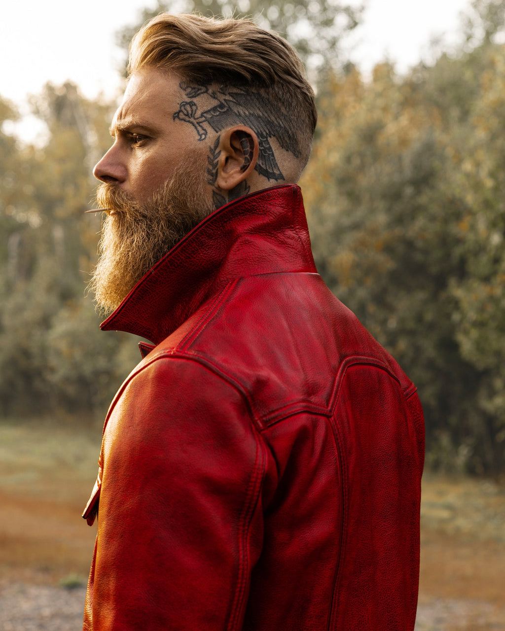 A man in profile wearing a bright red leather jacket, standing outdoors with trees blurred in the background.