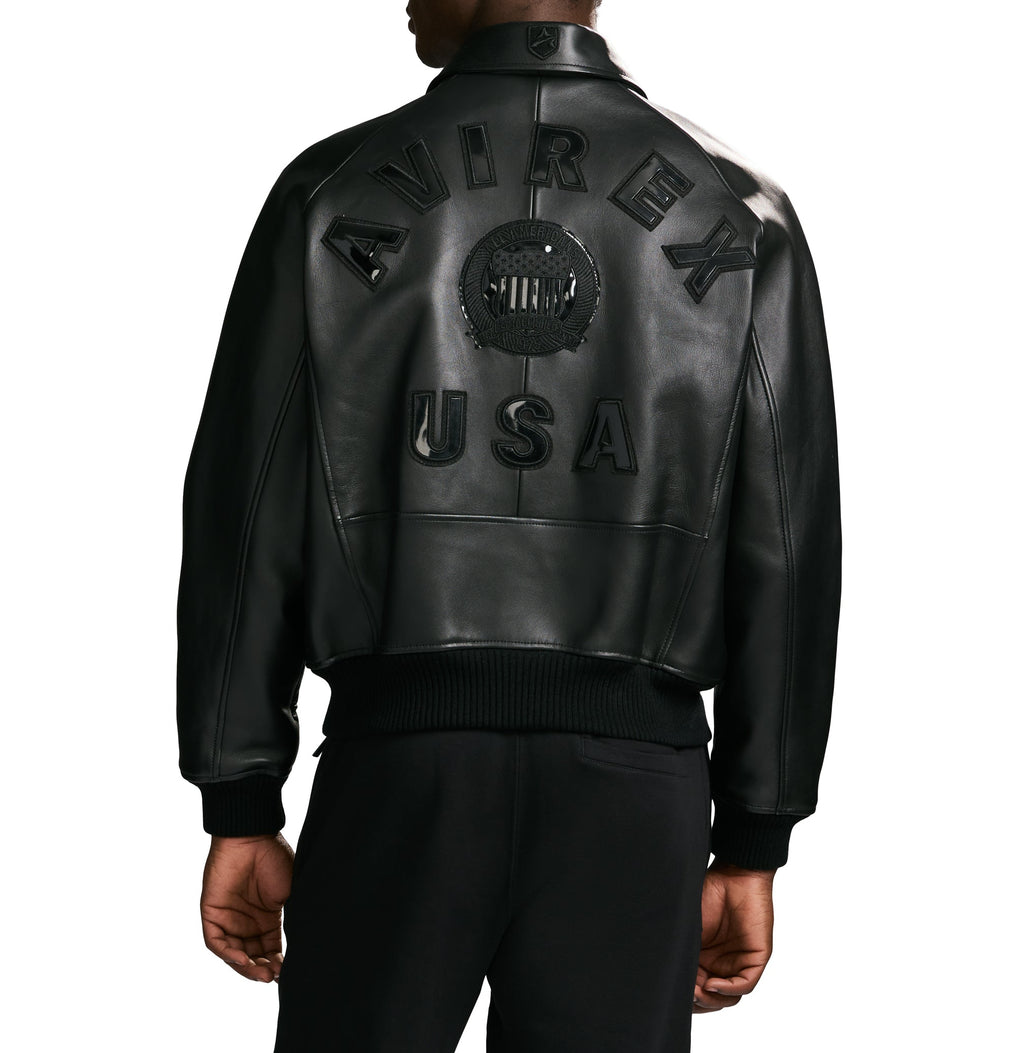 A person shown from the back wearing a black leather bomber jacket with large embossed 'AVIREX USA' lettering and a circular crest on the back, paired with black pants against a plain white studio background.