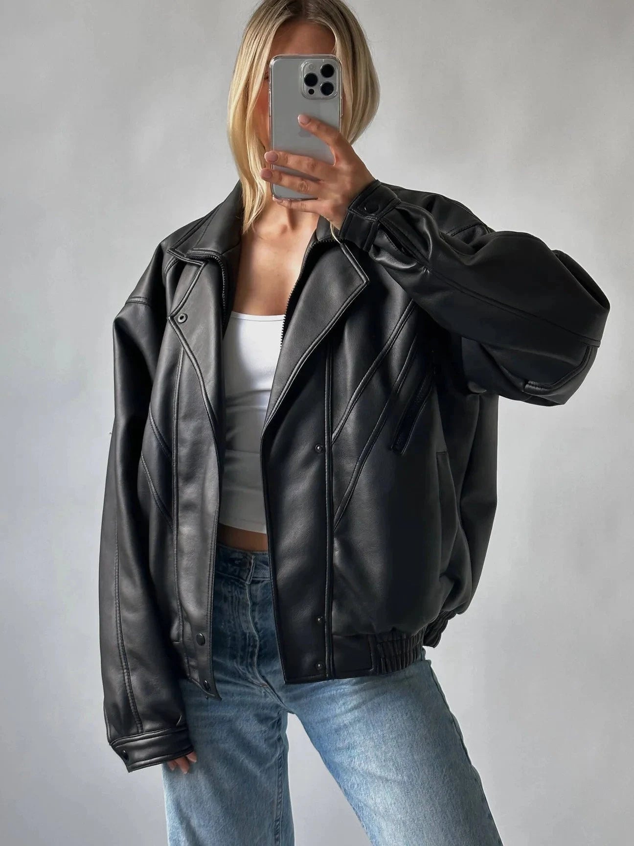 A woman takes a mirror selfie wearing a black faux leather moto jacket, a white crop top, and light blue jeans against a neutral gray background.