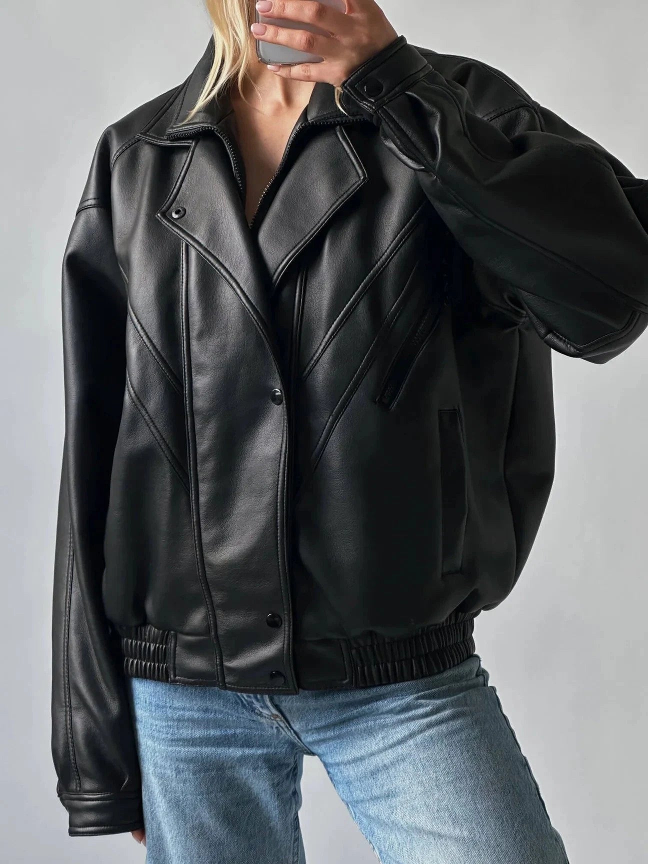A person wearing an oversized black leather jacket with diagonal paneling, ribbed knit trim at the hem and cuffs, and light blue jeans, photographed against a plain light gray studio background.