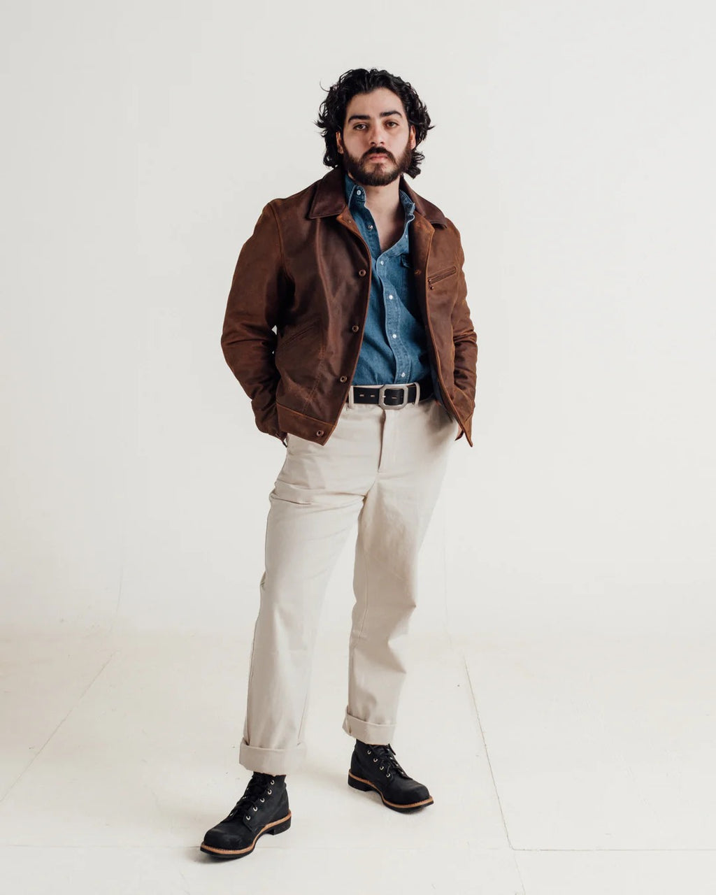 Man wearing a brown jacket, blue shirt, beige pants, and black boots on a white background