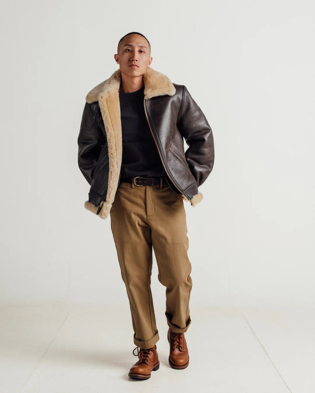 Man wearing a brown leather jacket with shearling lining, black shirt, tan pants, and brown boots on a white background
