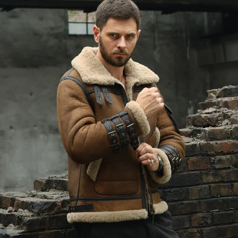 Man wearing a brown shearling jacket with leather arm guards standing in front of a brick wall.
