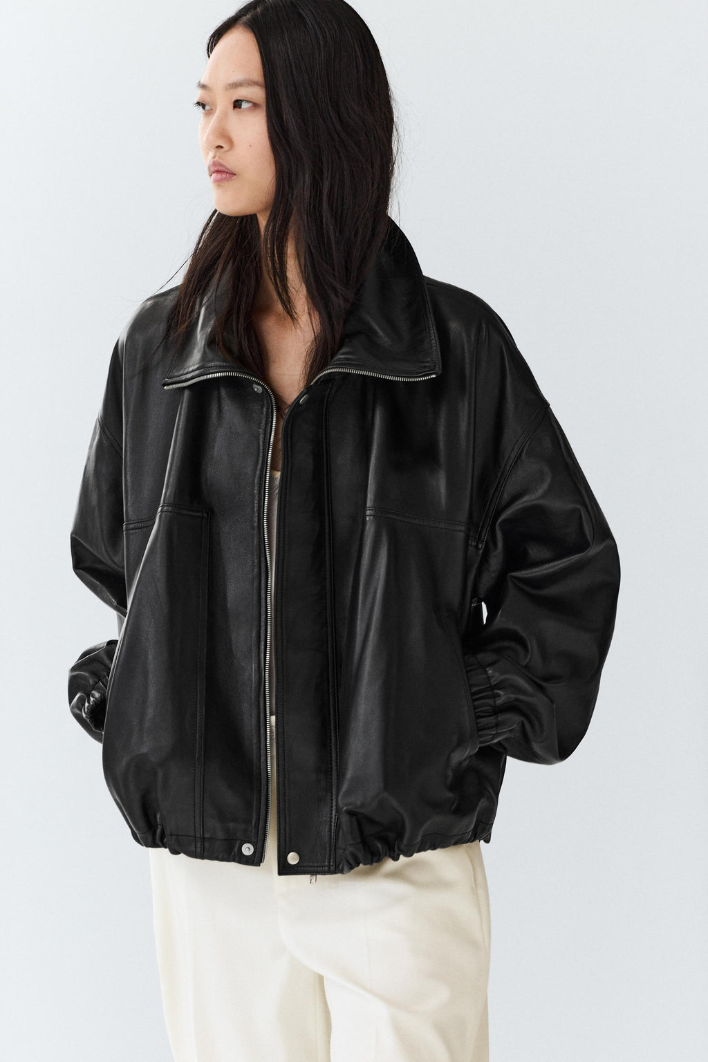 A woman wearing a black leather jacket with a high collar and front zipper, paired with cream-colored pants, photographed against a light gray studio background.