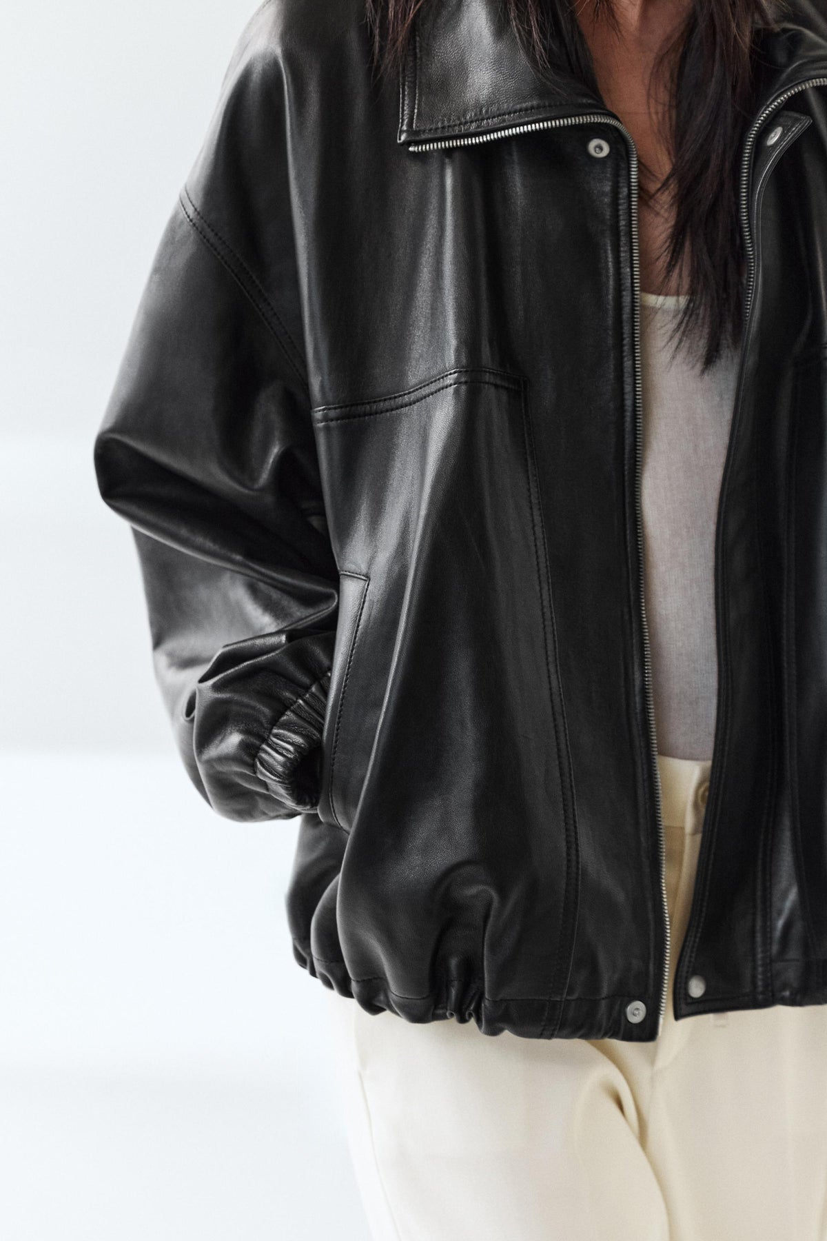 A close-up of a person wearing a black leather jacket over a light gray top and cream-colored pants, set against a clean white background.
