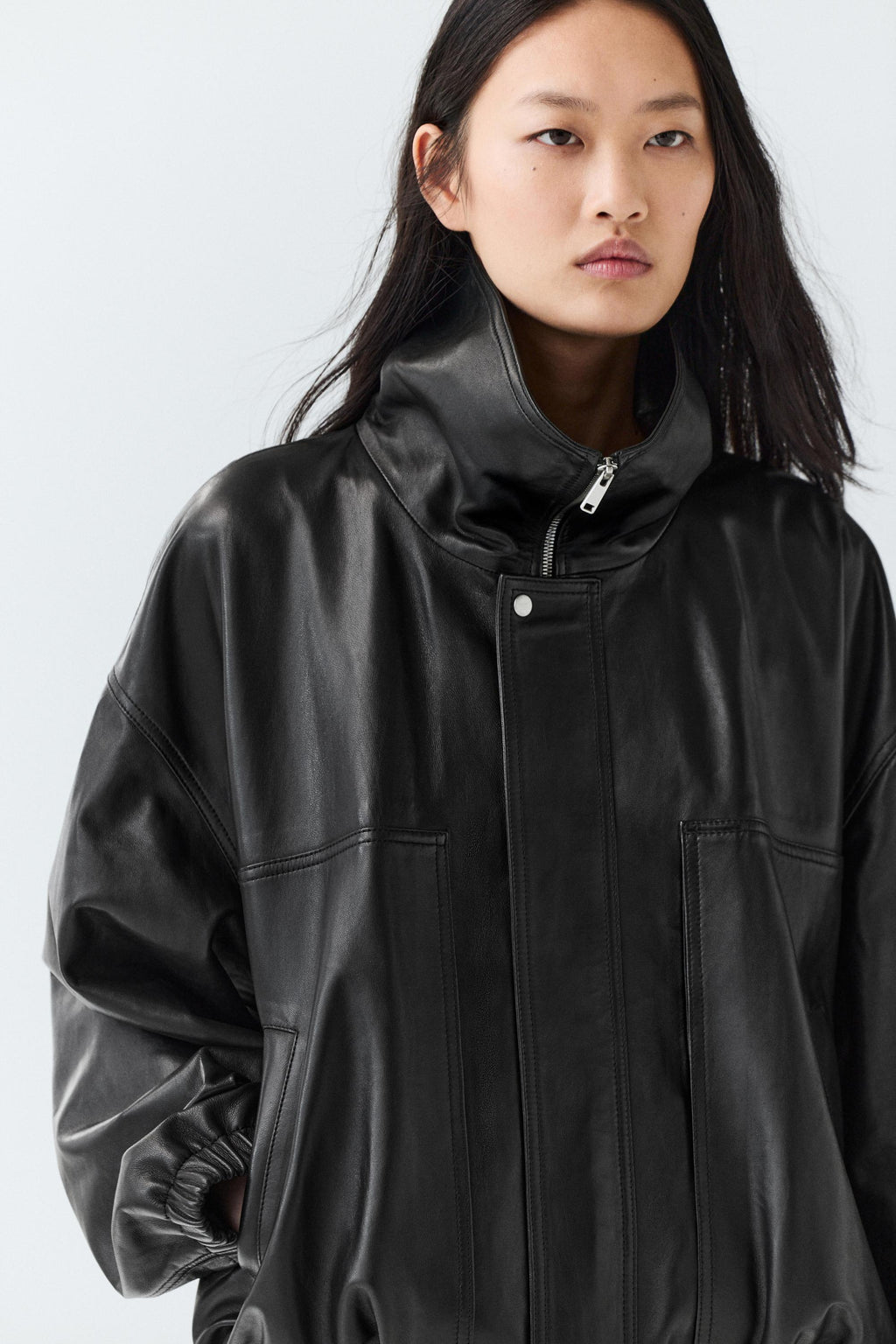 A model wearing a black leather jacket with a high collar and front zipper, photographed against a light gray studio background.