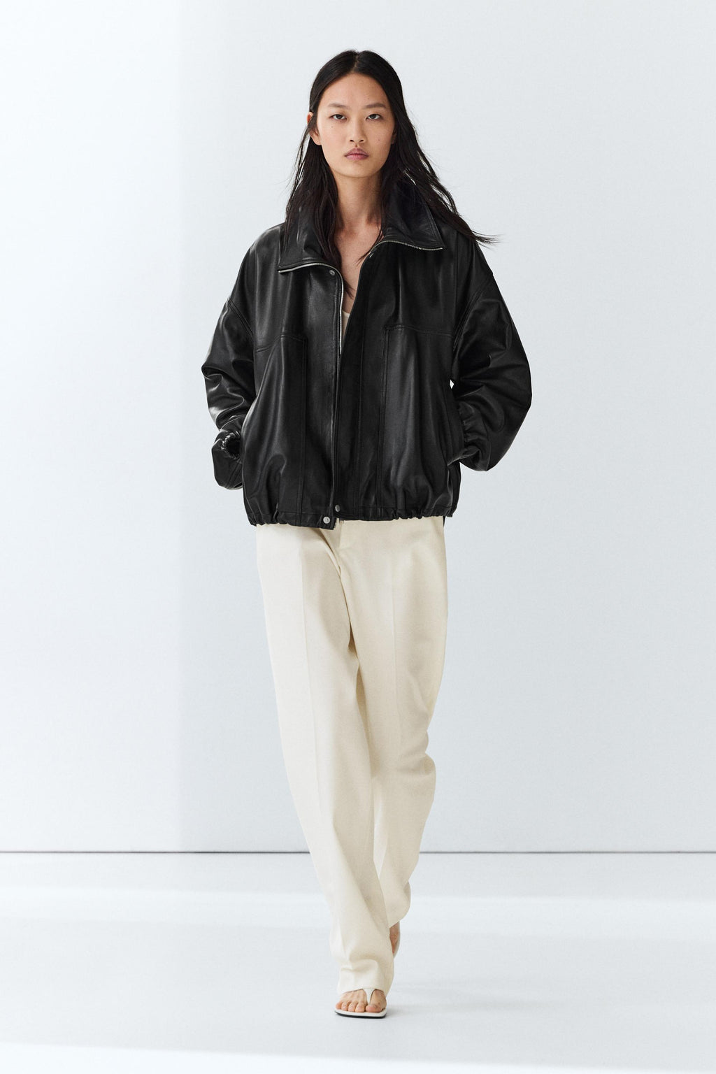A model wearing a black leather-look zip-up jacket and cream-colored wide-leg trousers, standing against a clean white studio background.