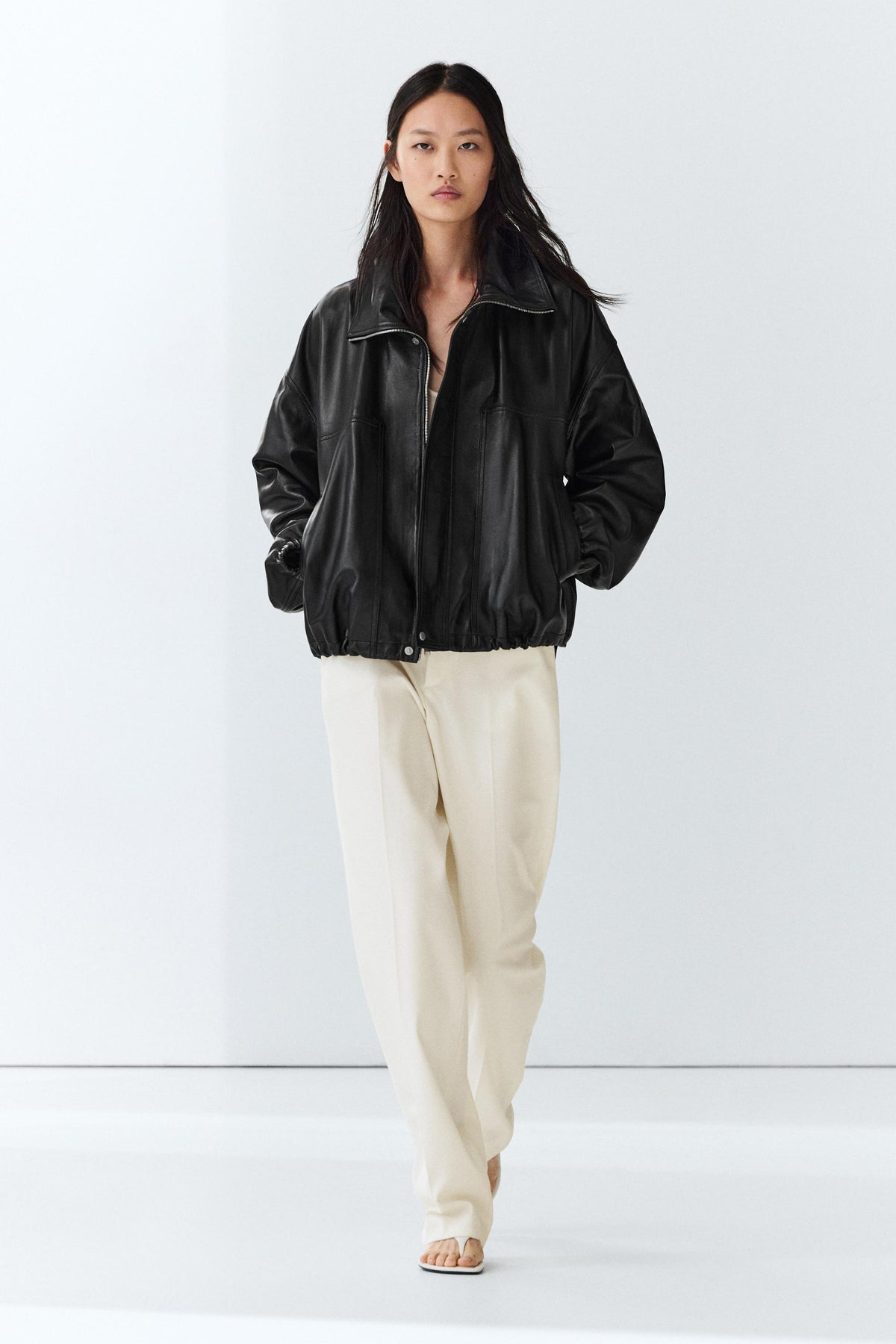 A model wearing a black leather-look zip-up jacket and cream-colored wide-leg trousers, standing against a clean white studio background.