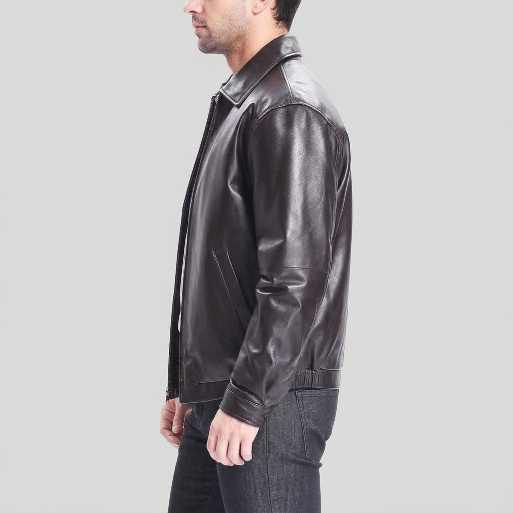 A man shown in profile wearing a dark brown leather jacket and dark jeans against a light gray studio background.